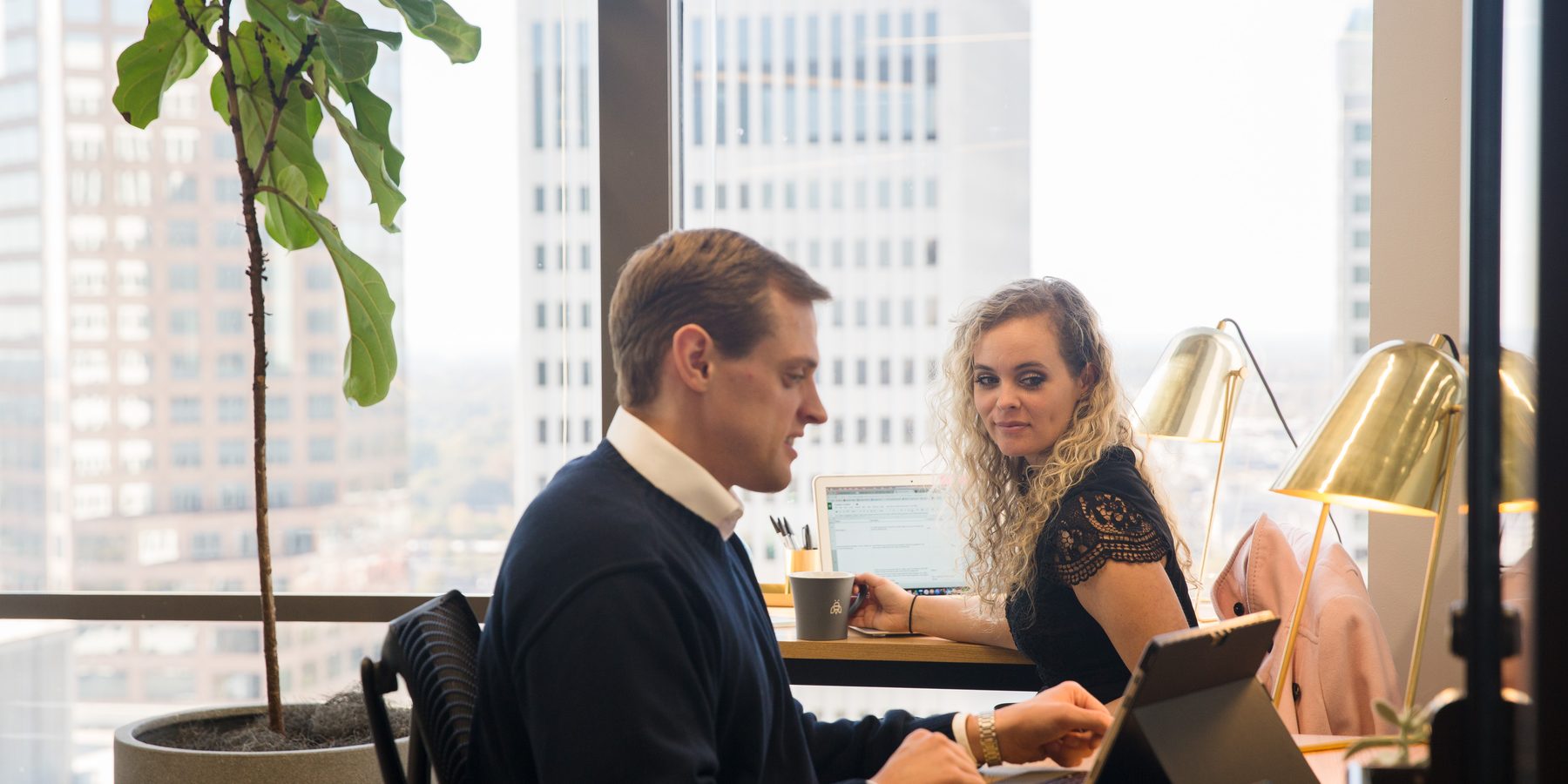 A man and woman working on laptops at a shared desk in a Charlotte coworking space, with the city skyline in the window