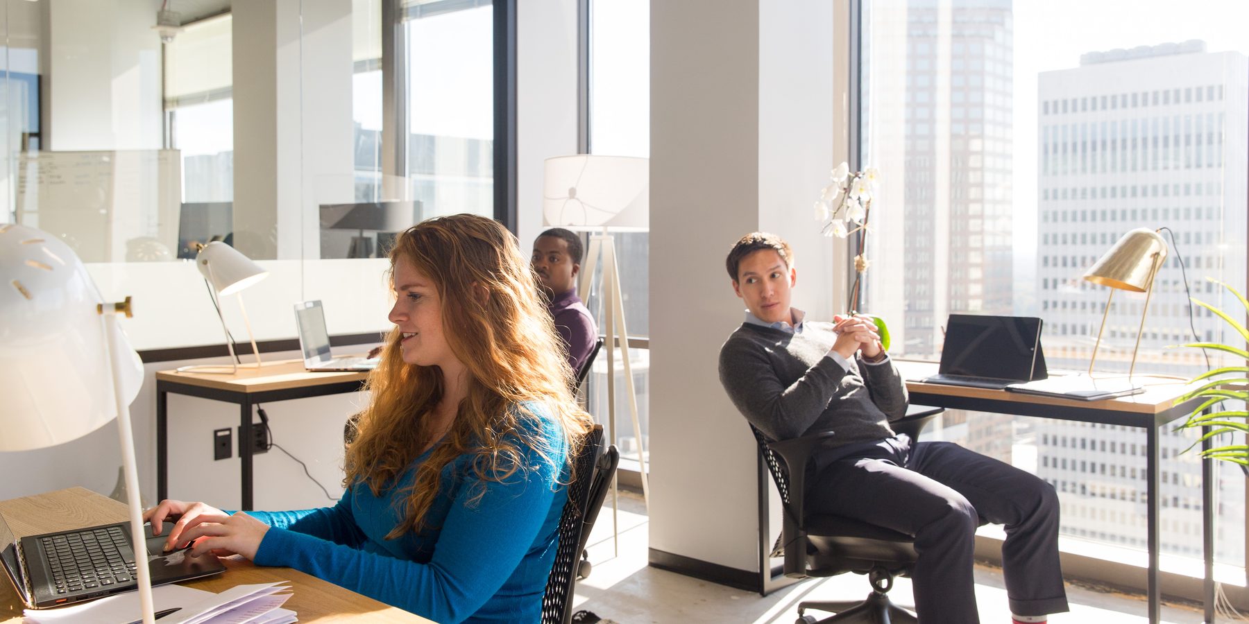 Three professionals working at desks in a bright Charlotte coworking office with the city skyline visible through large windows