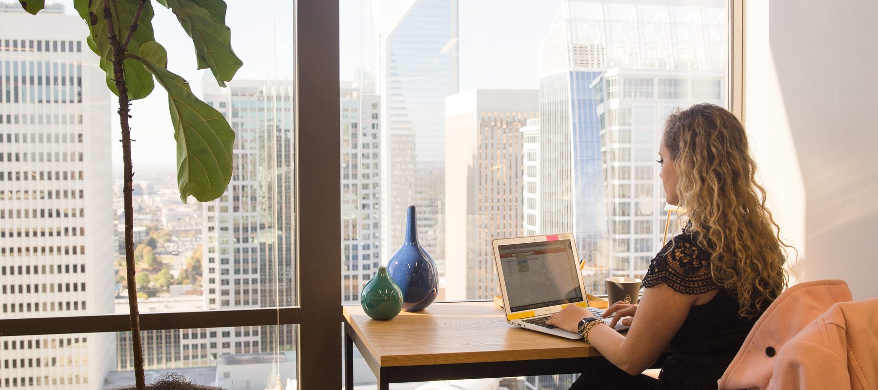 A woman working at a laptop in a Charlotte coworking space, with the Duke Energy Center and Uptown skyline visible through floor-to-ceiling windows