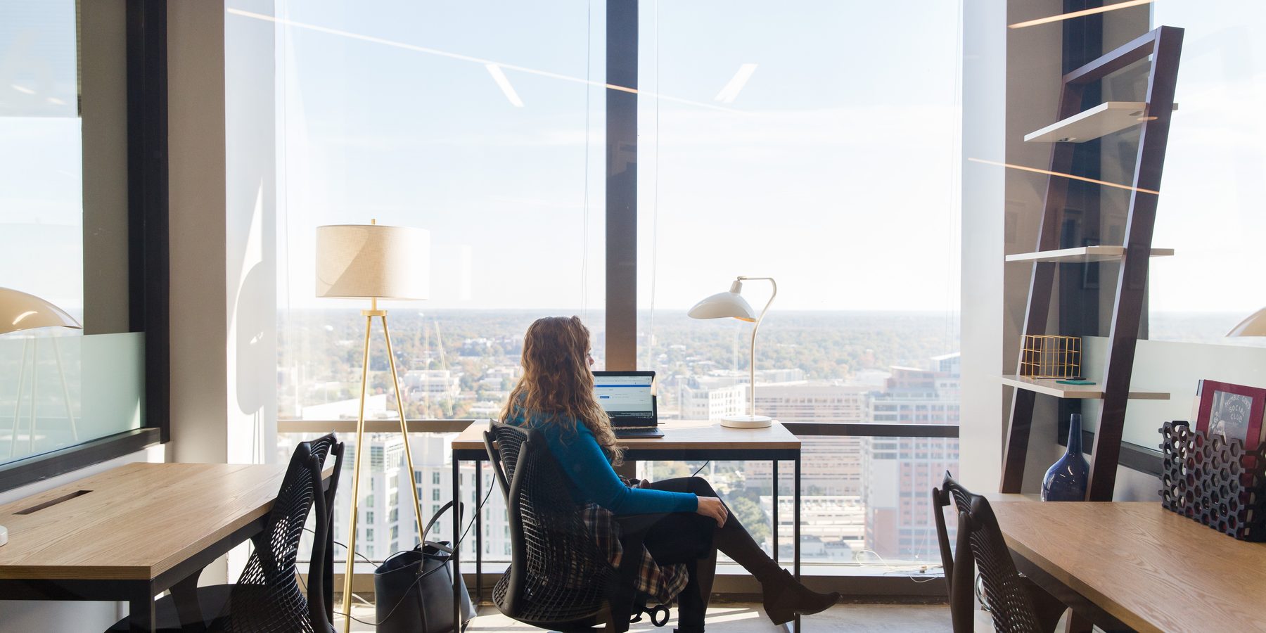 A woman working on a laptop at a desk facing the Charlotte skyline through floor-to-ceiling windows in a coworking office